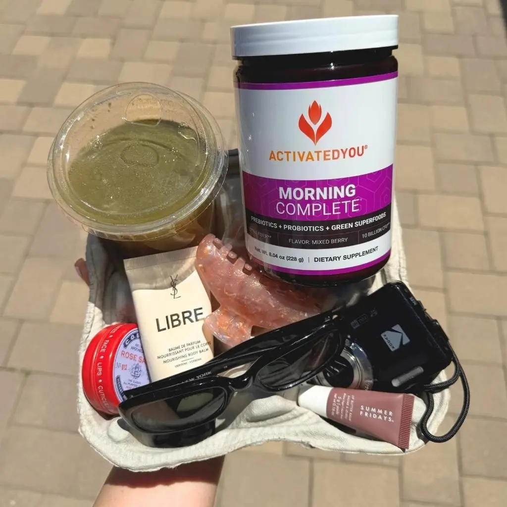 Someone carrying a four-cup carrier filled with sunglasses, a camera, a green beverage, and a jar of Morning Complete Mixed Berry